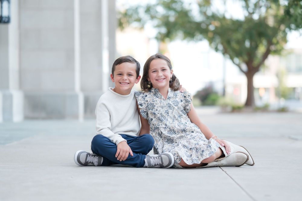 Two smiling children sit together outdoors on pavement, boy in white sweater and jeans, girl in floral dress.