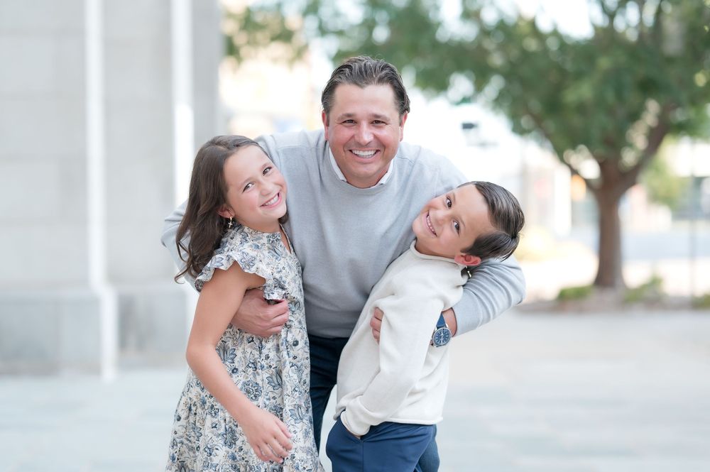A smiling man hugs two happy children outdoors in a warm family portrait photo.