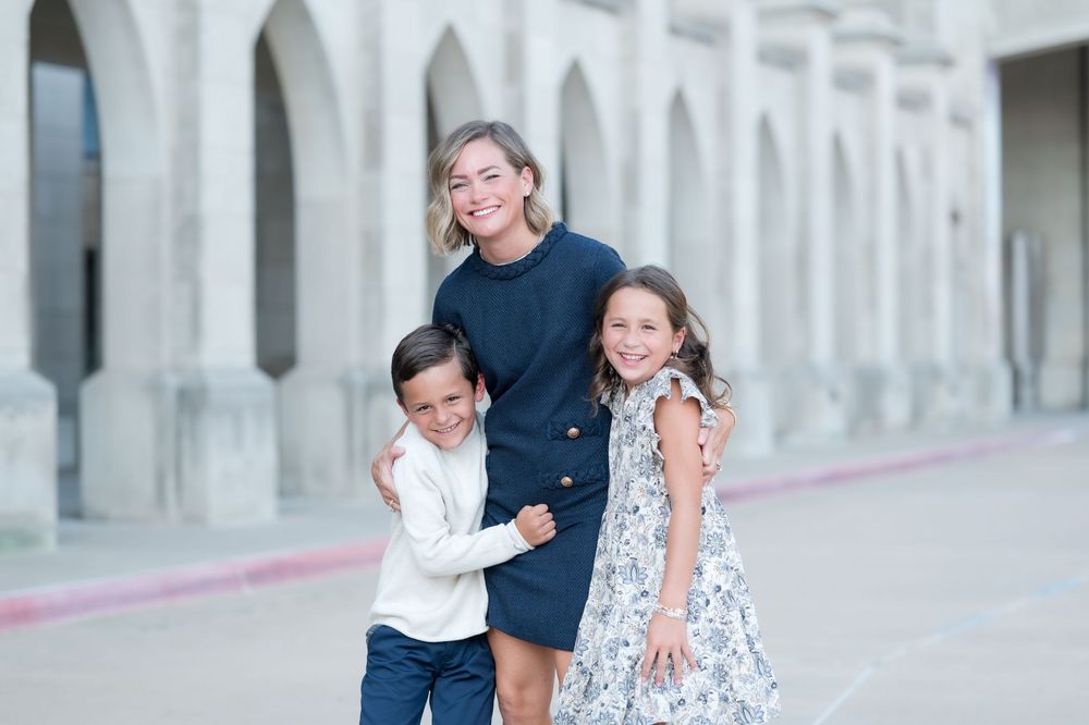 A smiling woman in a navy dress poses with two happy children in front of elegant arched columns outdoors.