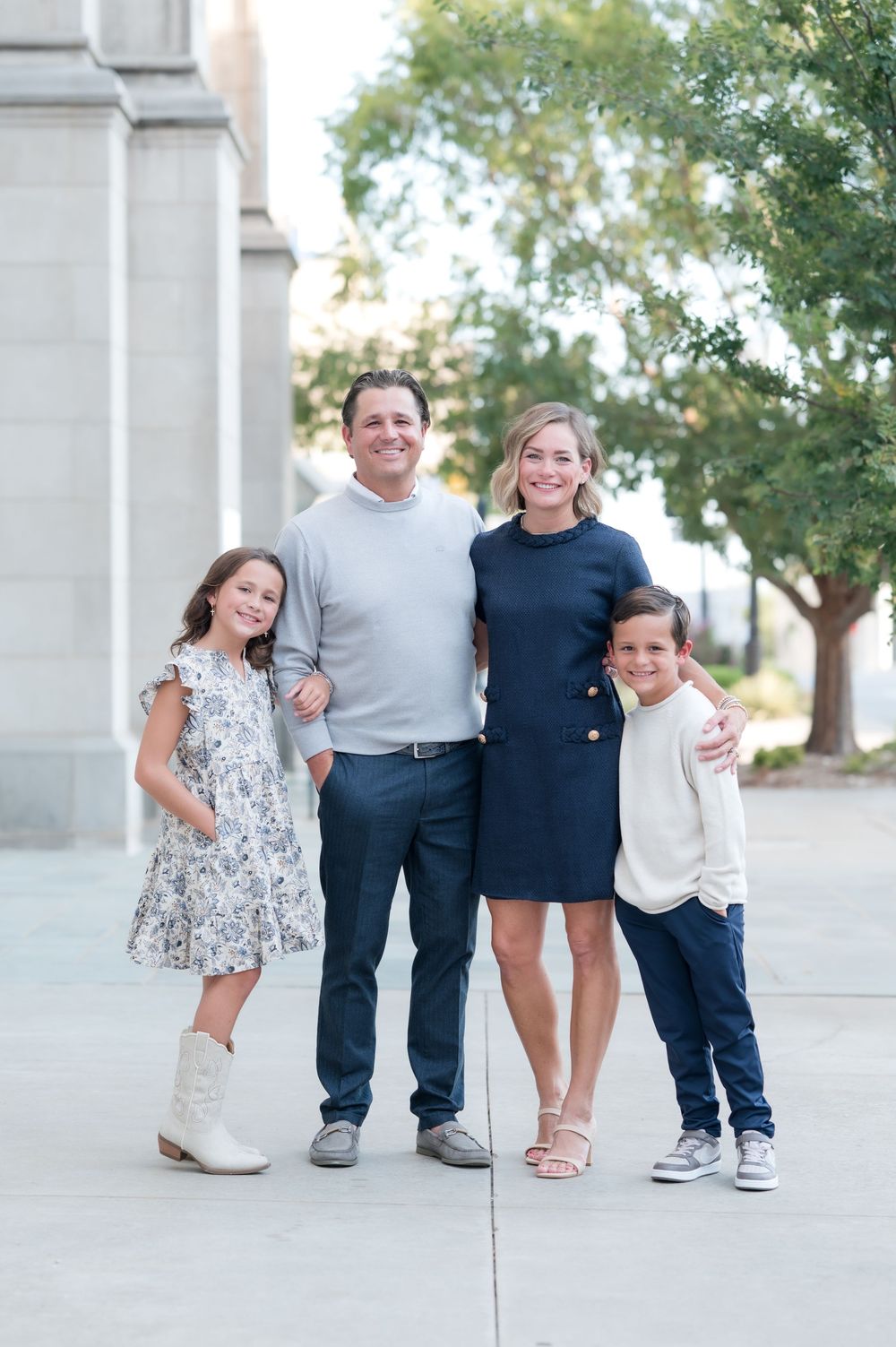 A smiling family of four poses outdoors, with two adults and two children dressed in coordinated navy and neutral outfits.