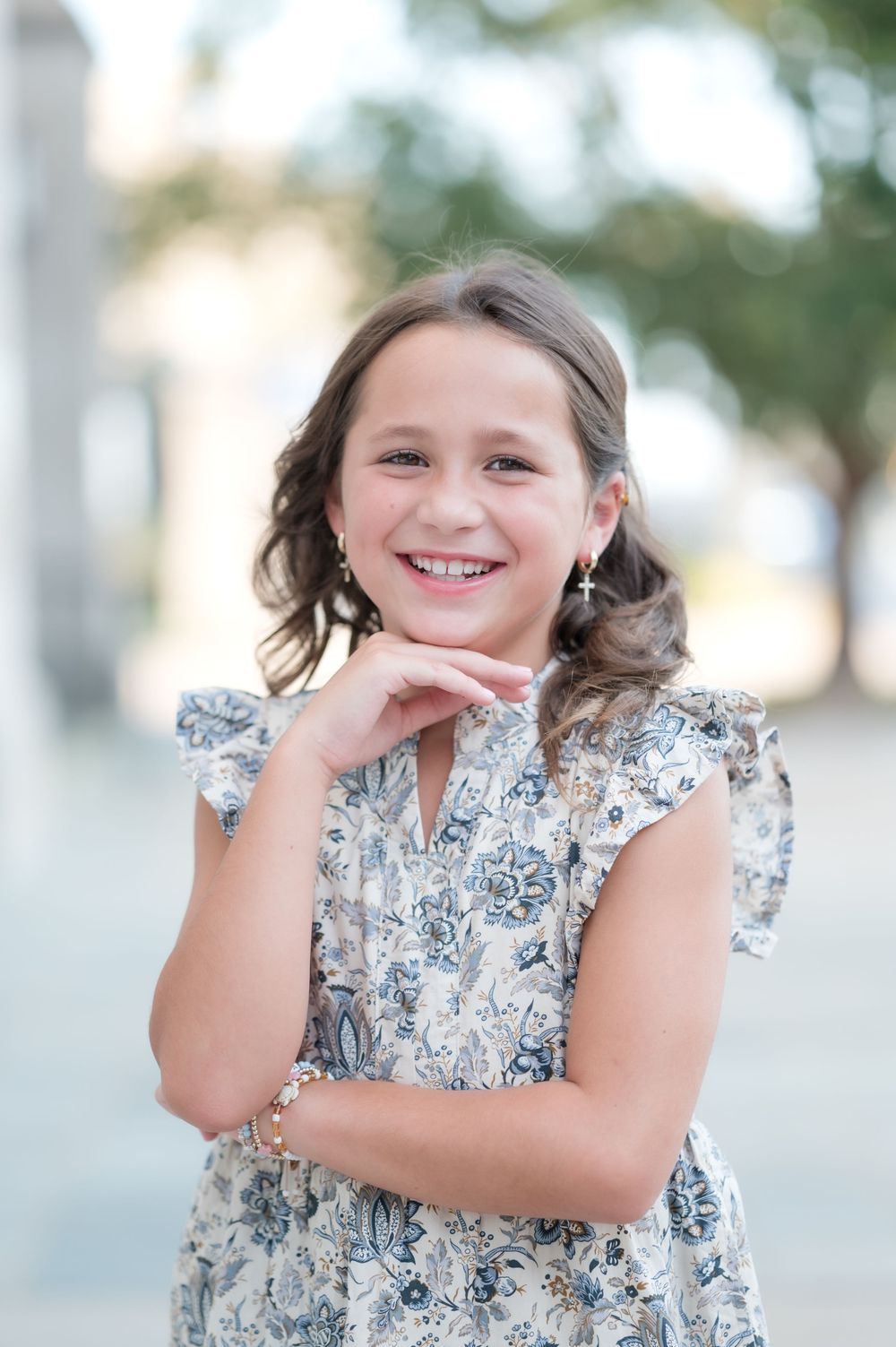 Smiling young girl in floral dress posing outdoors with hand under chin, wearing bracelet and earrings.