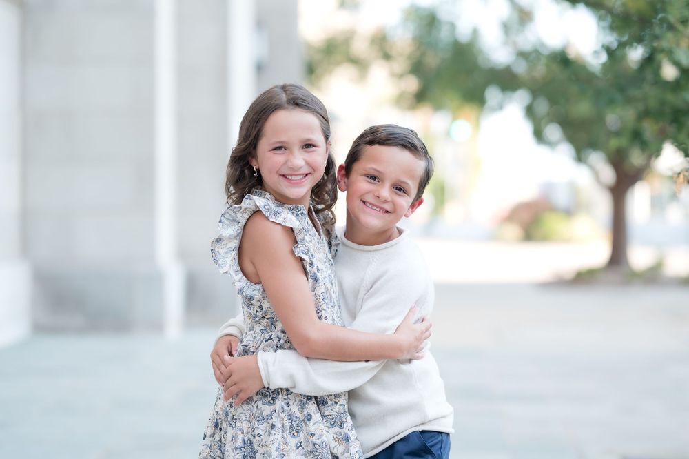 Two smiling children hugging outdoors, a girl in a floral dress and a boy in a white shirt, posing for a portrait.