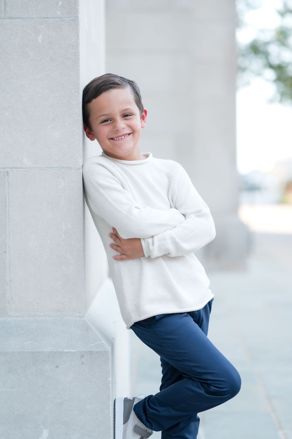 Smiling boy in white sweater and navy pants leaning against a pillar with arms crossed outdoors.