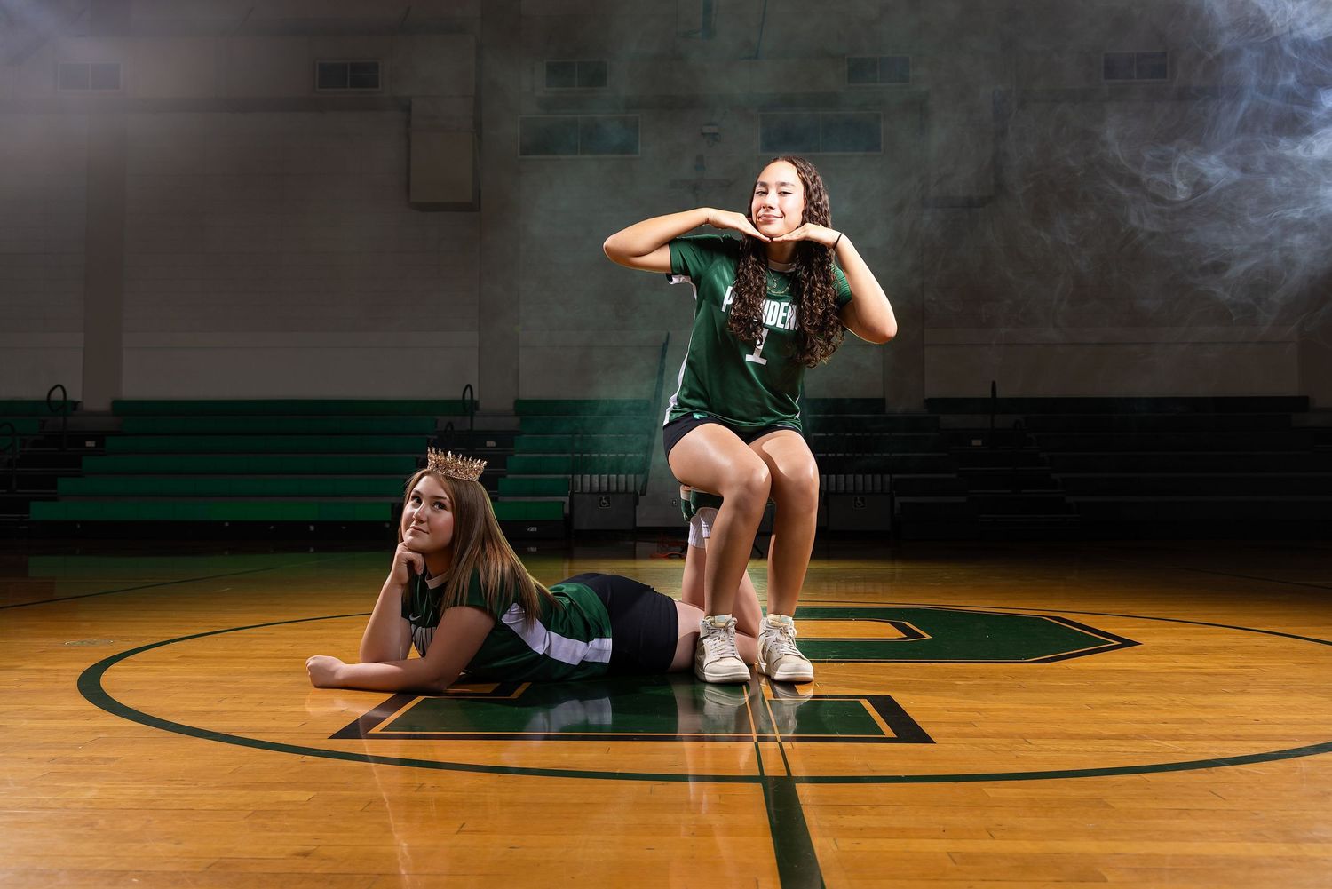Two female volleyball players pose on a gym court in green uniforms, one lying down and one crouching playfully.