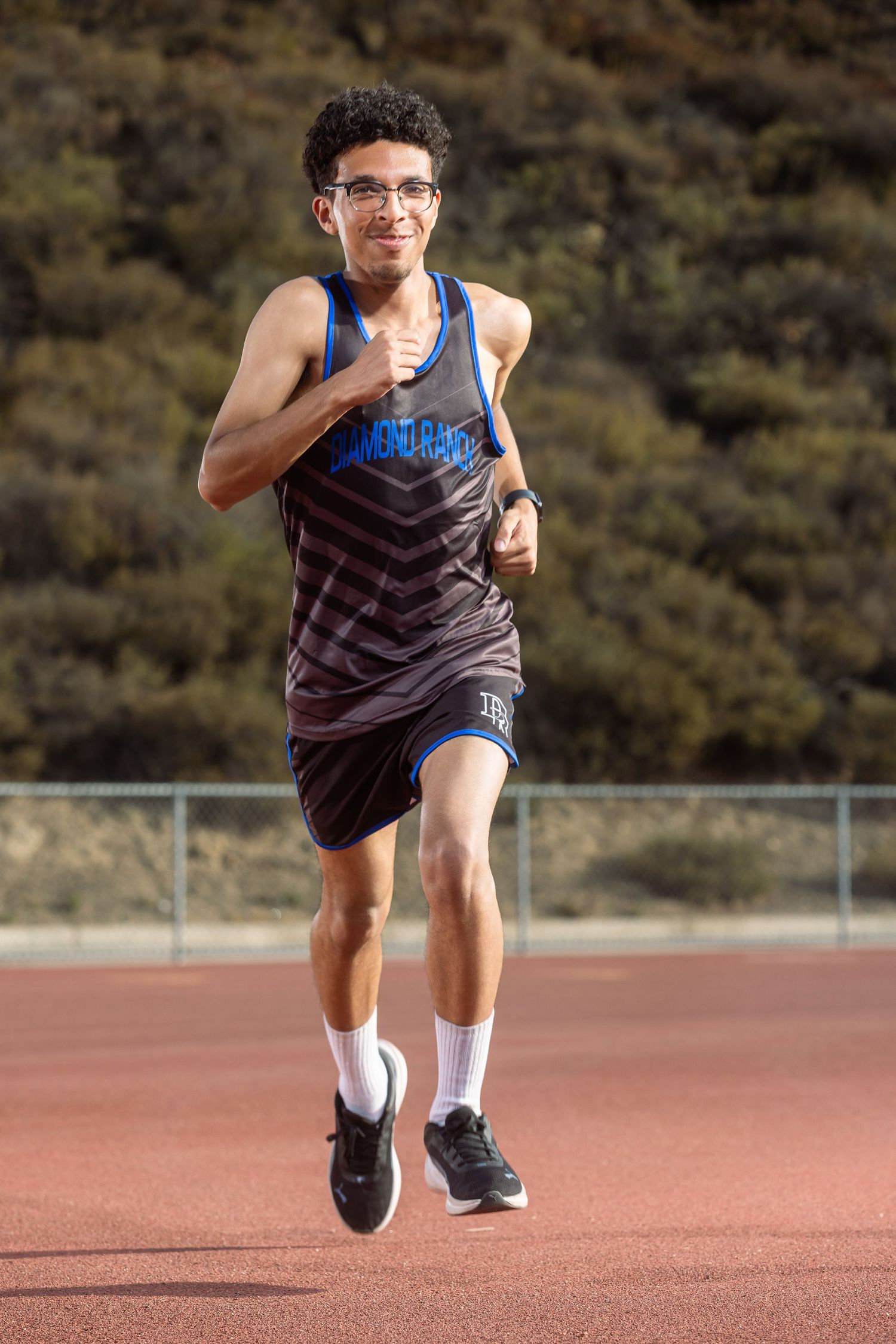 A young male athlete in track uniform runs on a red track with hills in the background.