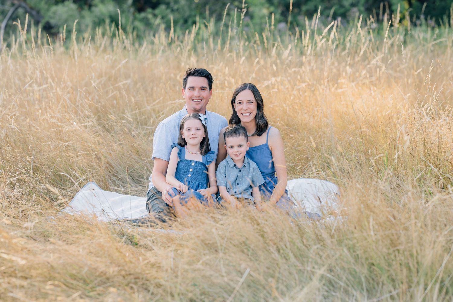 Family of four smiling together in a golden grass field, parents and two young children in denim outfits.