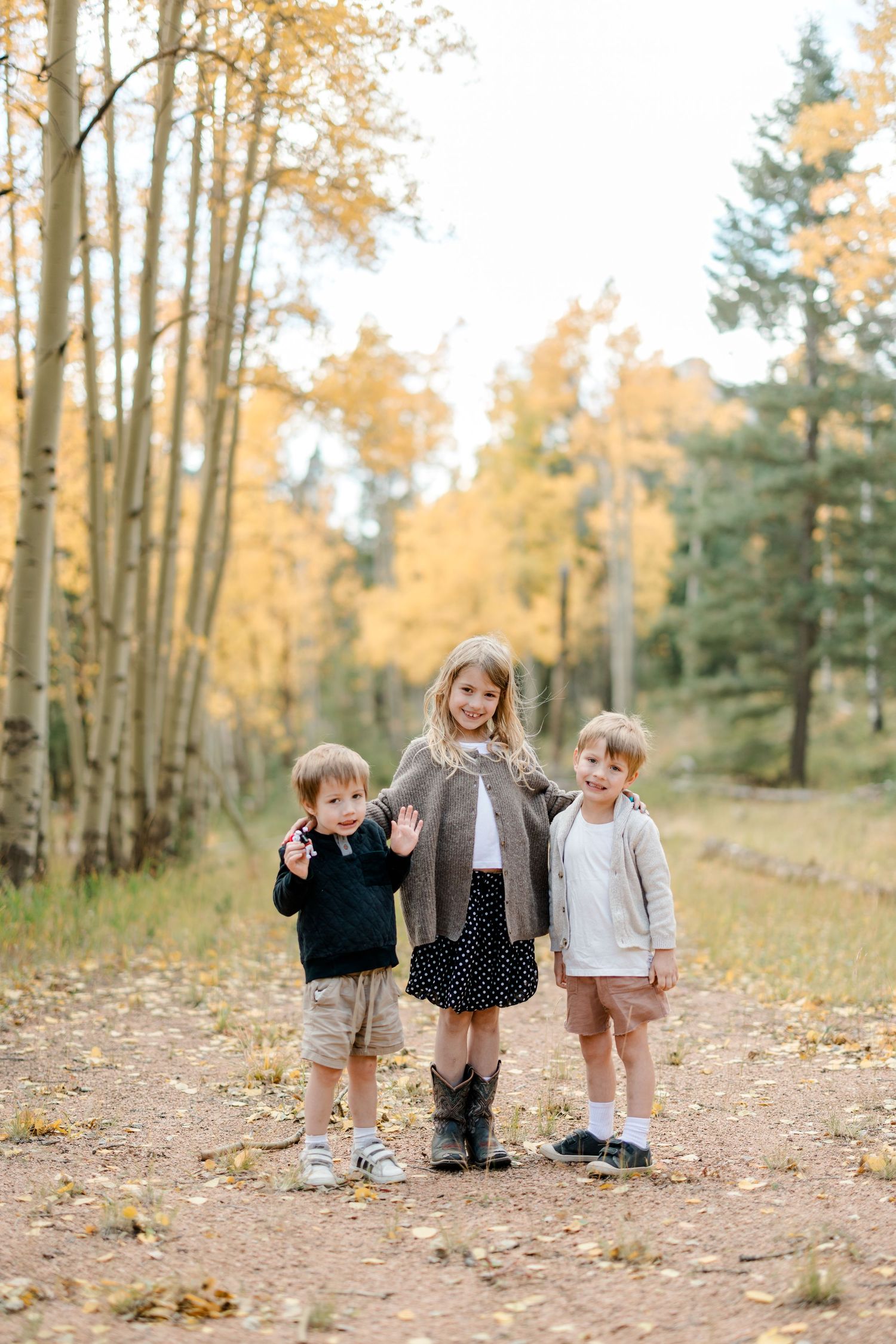 Three children pose on a dirt path surrounded by golden autumn aspen trees in a forest setting.