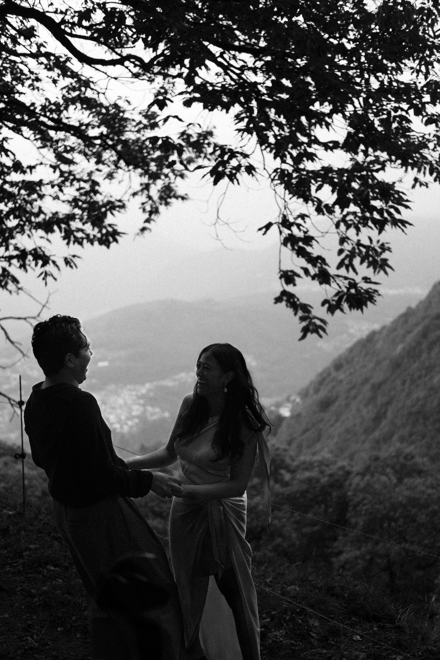Black and white photo of a couple holding hands on a misty mountain overlook framed by tree branches.