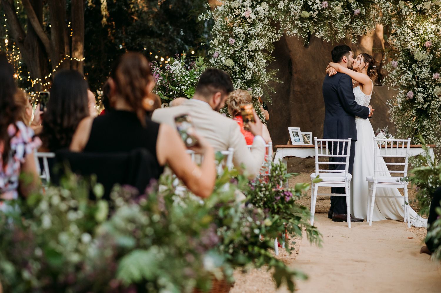 Couple sharing first dance at outdoor wedding ceremony surrounded by lush greenery and fairy lights.