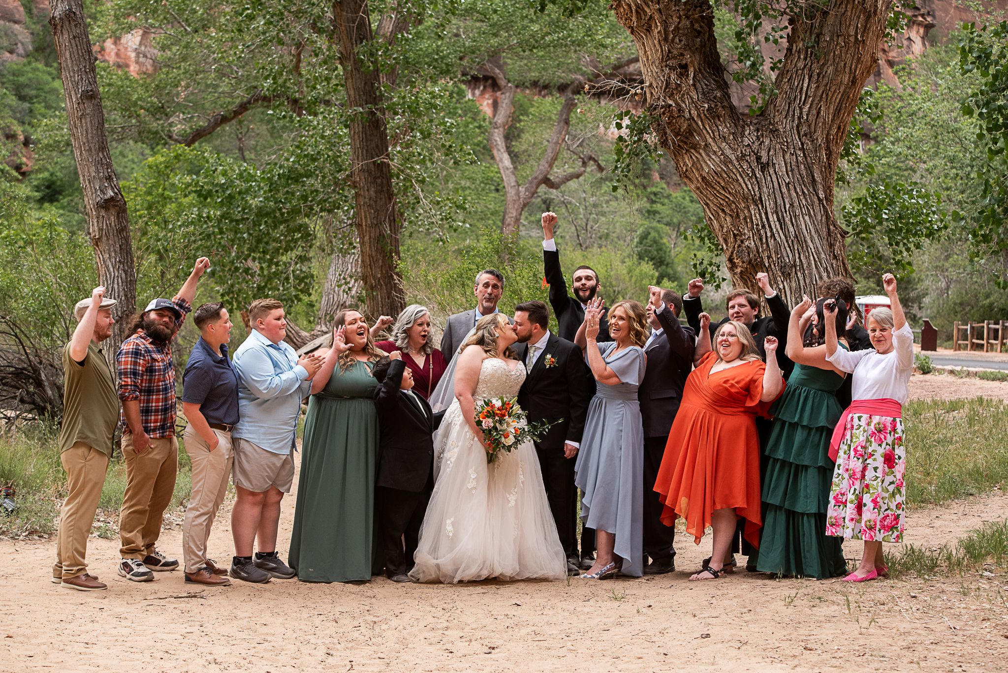 Wedding party celebrating outdoors near trees, bride and groom surrounded by cheering guests in colorful attire.