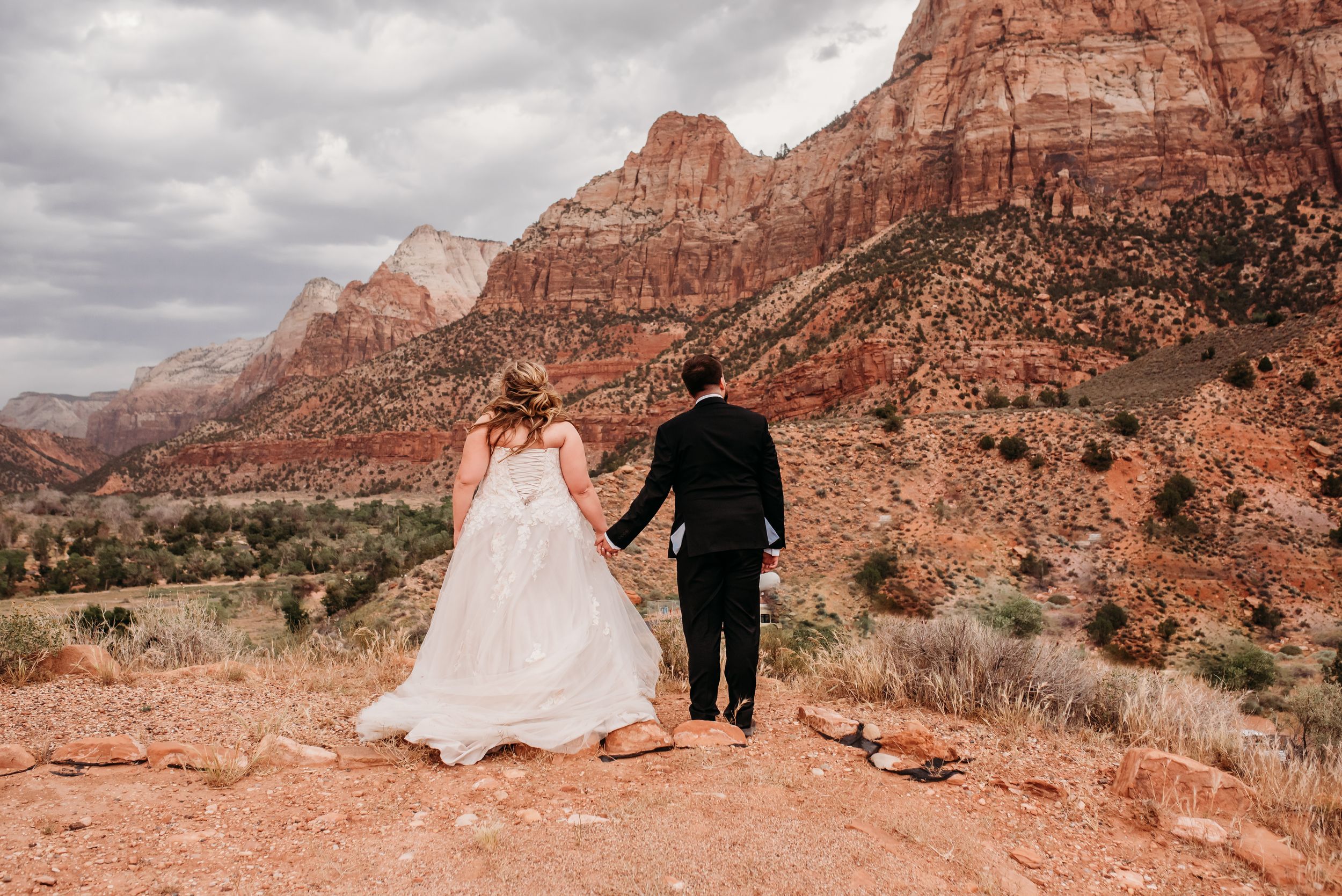 Bride and groom hold hands overlooking stunning red rock canyon landscape at Zion National Park on their wedding day.