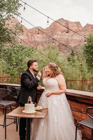 Bride and groom cutting wedding cake on outdoor patio with stunning red rock Zion National Park mountains backdrop.