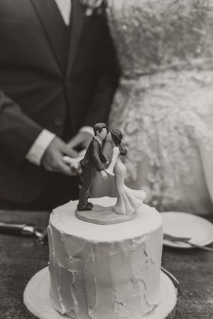 Black and white photo of a wedding cake topper featuring a kissing couple, with bride and groom standing behind the white frosted cake.