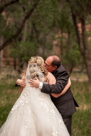 Bride and groom share a tender embrace outdoors, with lace wedding gown and veil, surrounded by lush greenery.