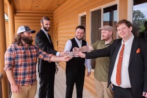Five men in semi-formal attire celebrate together on a wooden cabin porch, joining hands in a group toast.