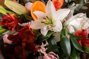 Close-up of a bridal bouquet featuring white lilies, red alstroemeria, orange roses, and a sparkling diamond engagement ring.