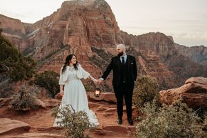Couple in wedding attire holding hands at Zion National Park with red rock canyon mountains backdrop.