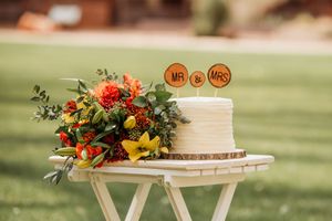 White wedding cake with Mr & Mrs topper and vibrant orange floral arrangement on rustic wood slice table outdoors.