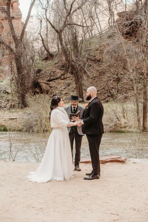 Couple exchanges vows outdoors near a river with an officiant, surrounded by bare trees and red rock cliffs.