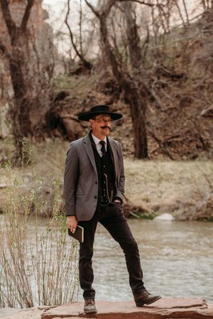 Man in cowboy hat, mustache, gray blazer, and black pants stands by a river in a wooded canyon setting.