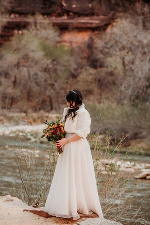 Bride in white gown holding colorful bouquet stands by river with red rock canyon backdrop.