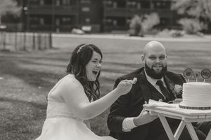 Black and white photo of a laughing bride feeding wedding cake to her bearded groom outdoors.