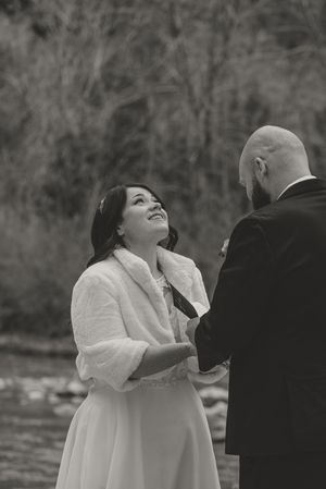 Black and white photo of a smiling bride in a fur shawl holding hands with a groom during an outdoor wedding ceremony.