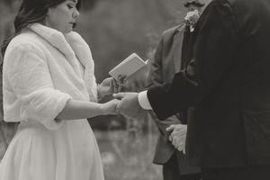 Black and white wedding ceremony photo of bride in white fur wrap exchanging rings with groom outdoors.