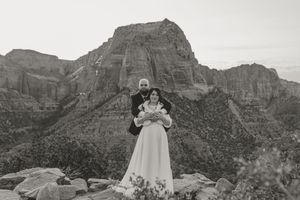 Couple poses in wedding attire on rocky terrain with dramatic mountain cliffs in black and white photo.