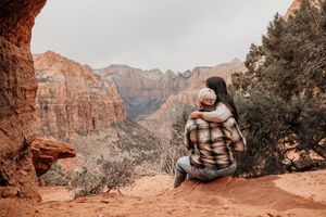 A couple sits embracing on a red rock ledge overlooking the stunning canyon landscape of Zion National Park.