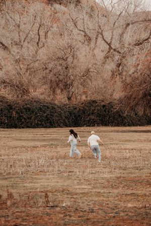 Couple running hand-in-hand through a dry autumn field with bare trees and rocky hillside backdrop.
