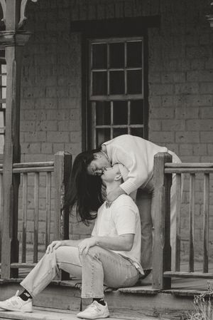 Black and white photo of a couple sharing a romantic kiss on a rustic wooden porch steps of an old house.