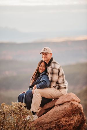 Couple sitting on a red rock cliff embracing, with scenic canyon views and soft sunset light behind them.