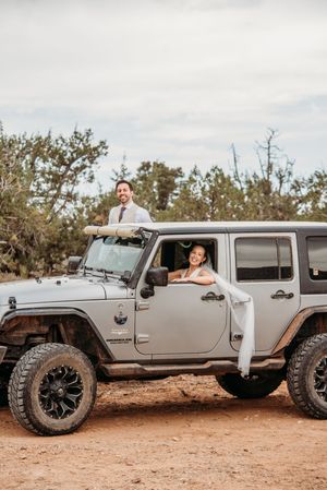 Bride and groom posing with a gray Jeep Wrangler on a dirt trail, surrounded by desert shrubs.