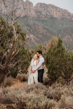 Couple in wedding attire embraces in desert landscape with juniper trees and red rock mountains in background.
