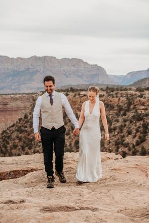 A couple in wedding attire holds hands on a rocky overlook with red rock canyon views in the background.