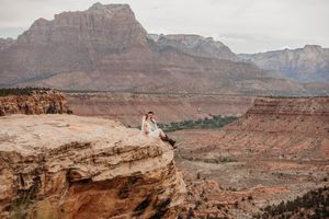 Couple in wedding attire sitting on a rocky cliff edge overlooking Zion National Park red rock canyon landscape.