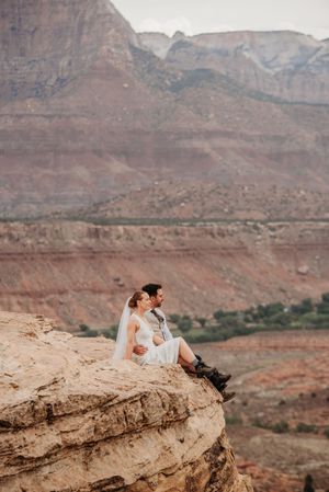 Bride and groom sit on a rocky cliff edge with stunning red canyon mountains backdrop during their elopement.