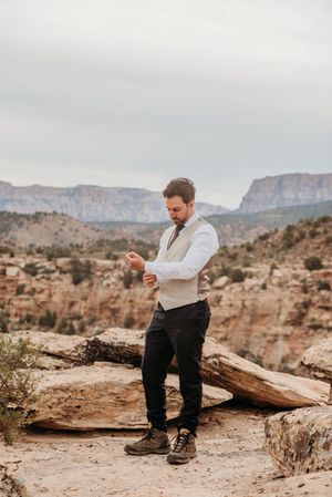 Groom in vest and tie adjusting cufflinks on rocky overlook with canyon mountains backdrop.