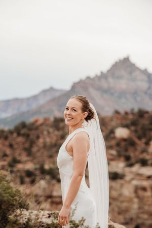 Smiling bride in white gown and veil poses outdoors with dramatic red rock mountain scenery behind her.