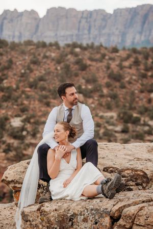 Bride and groom sit on rocky outcrop with red canyon mountains backdrop during outdoor desert wedding photo session.