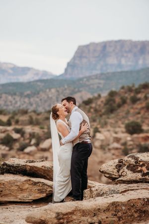 Bride and groom embrace laughing on rocky desert landscape with mountain cliffs backdrop during outdoor wedding ceremony.