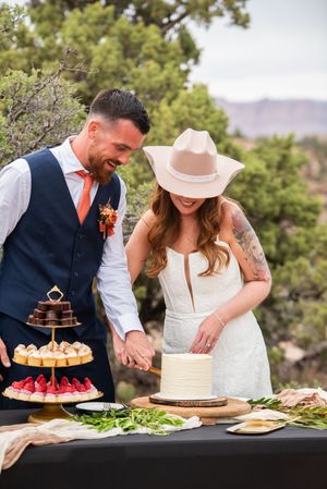 Bride in cowboy hat and groom cutting wedding cake outdoors at rustic desert wedding with tiered dessert display.