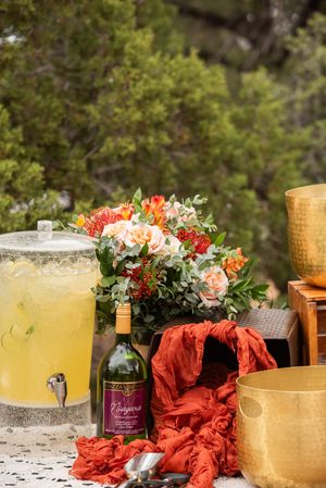 Outdoor beverage station with lemonade dispenser, wine bottle, coral floral arrangement, and gold accents on lace tablecloth.