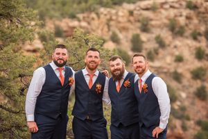 Four groomsmen in navy vests and orange ties pose outdoors with rocky mountain scenery behind them.