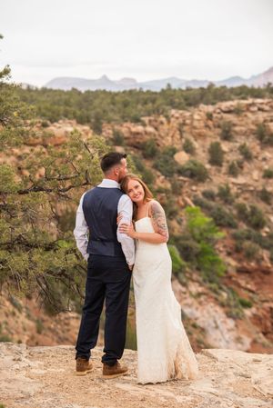 Bride and groom embrace on rocky overlook with red canyon landscape backdrop during outdoor desert wedding ceremony.