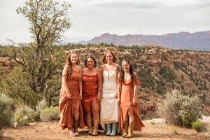 Four women in rust and cream dresses pose outdoors with red rock canyon landscape in background.