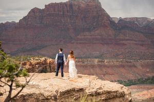Couple in wedding attire standing on red rock cliff overlooking Zion National Park canyon landscape.