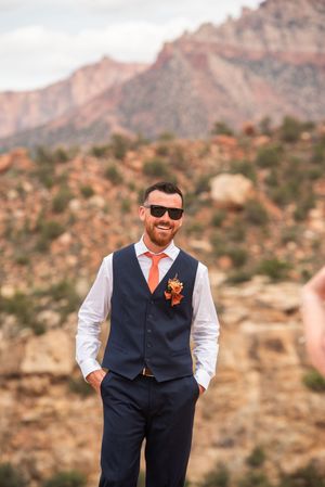Smiling groom in navy vest, orange tie, and sunglasses stands outdoors with red rock mountains in the background.