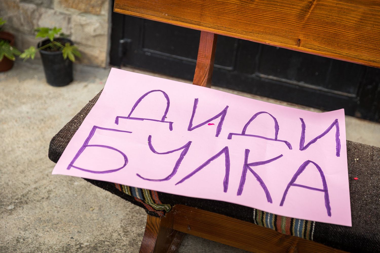 Pink paper sign with purple Cyrillic text reading ДИДИ БУЛКА placed on a wooden bench outdoors.